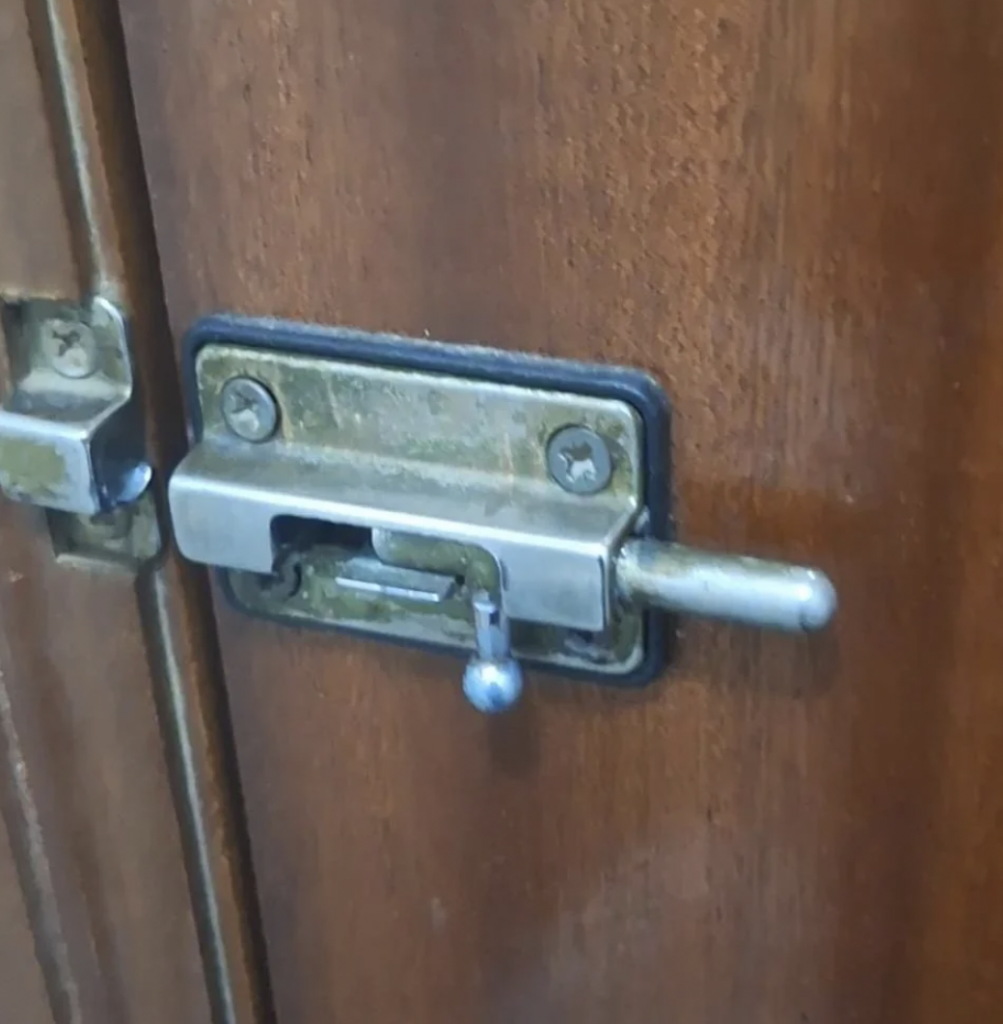 A close-up image of a brass sliding bolt latch on a wooden door. The latch is secured with visible screws and has a small handle for sliding. The door appears to be made of dark brown wood.