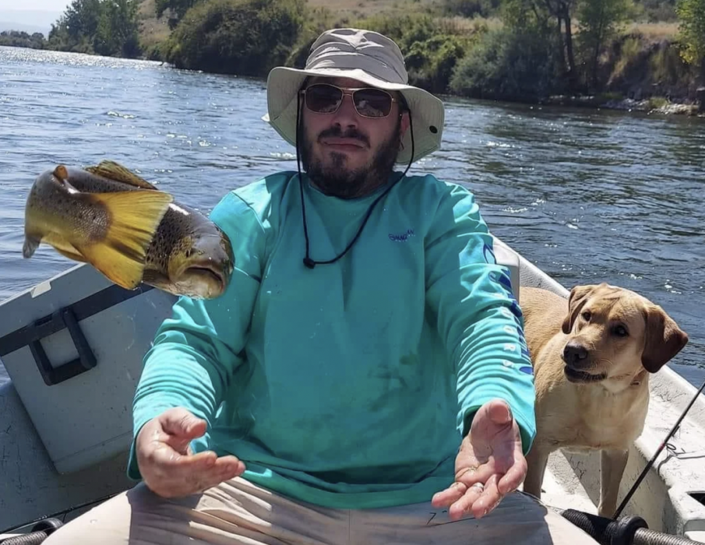 A man sitting in a boat on a river, wearing a large-brimmed hat and sunglasses, is tossing a fish in the air with his hands. A dog with a light brown coat is sitting beside him, looking at the fish. Greenery lines the riverbank in the background.