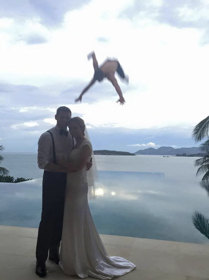 A couple in wedding attire pose together, standing close and holding each other in front of a scenic infinity pool with a backdrop of the ocean and mountains. A person is captured mid-air doing a backflip or some sort of acrobatic move in the background.