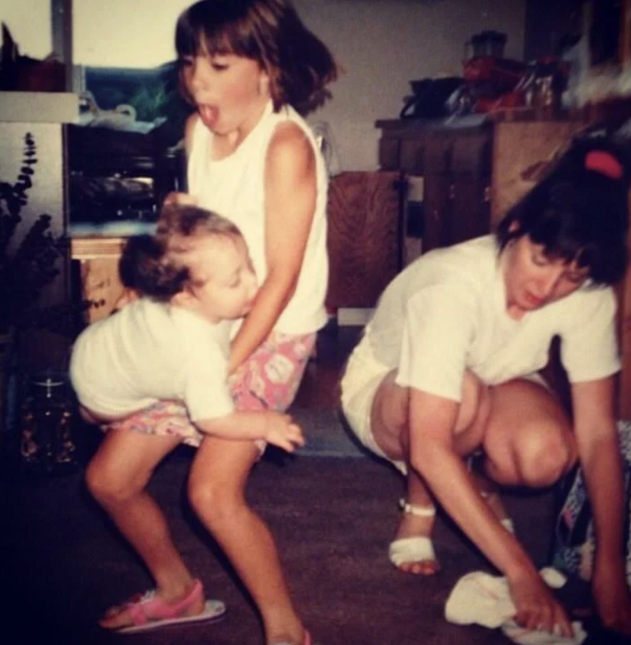 Three children are indoors. One child is carrying a younger child who appears to be trying to climb on them. The third child is crouched on the floor, possibly cleaning up a spill. The scene captures a moment of playful chaos.