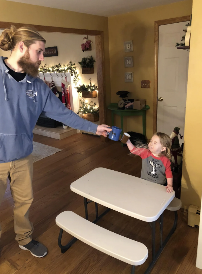 A man with a beard, wearing a blue hoodie, is handing a blue cup to a young child seated at a small white table with matching benches. The child, wearing a red and gray shirt, reaches out to accept the cup. Holiday decorations are visible in the background.