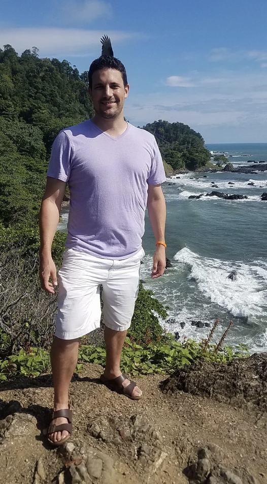 A man in a light purple t-shirt and white shorts stands on a cliff overlooking a rocky coastline with waves crashing against the shore. The sky is clear with a few clouds, and dense greenery surrounds the area. He is smiling and standing on a dirt path.