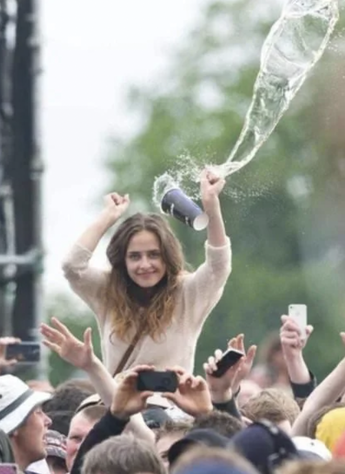 A woman stands above a crowd at an outdoor event, appearing to joyfully throw a drink from a cup. The liquid arcs through the air, captured mid-splash. People around her are taking photos and raising their hands. She has long hair and is wearing a light-colored sweater.