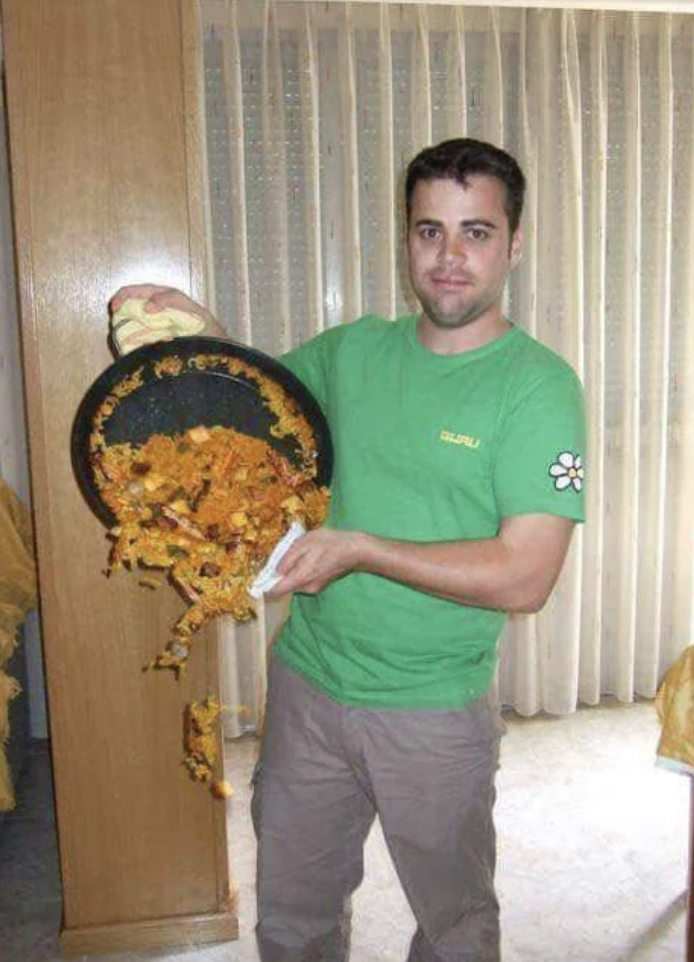 A man in a green t-shirt and beige pants is holding a pan, spilling food from it onto the floor. He appears to be in a kitchen with light-colored curtains in the background. He looks at the camera with a neutral expression.