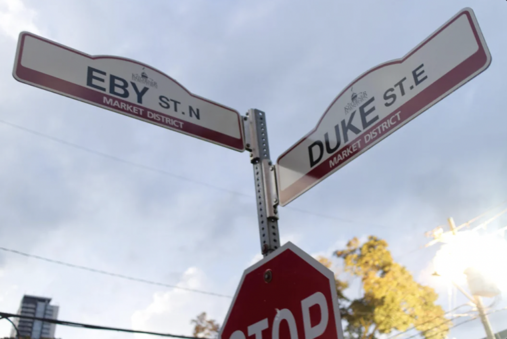 Street signs for Eby St. N and Duke St. E are mounted on a pole in the market district, with a stop sign at the bottom. The sky is overcast, with some clouds and power lines visible in the background. Trees and buildings can be seen in the distance.