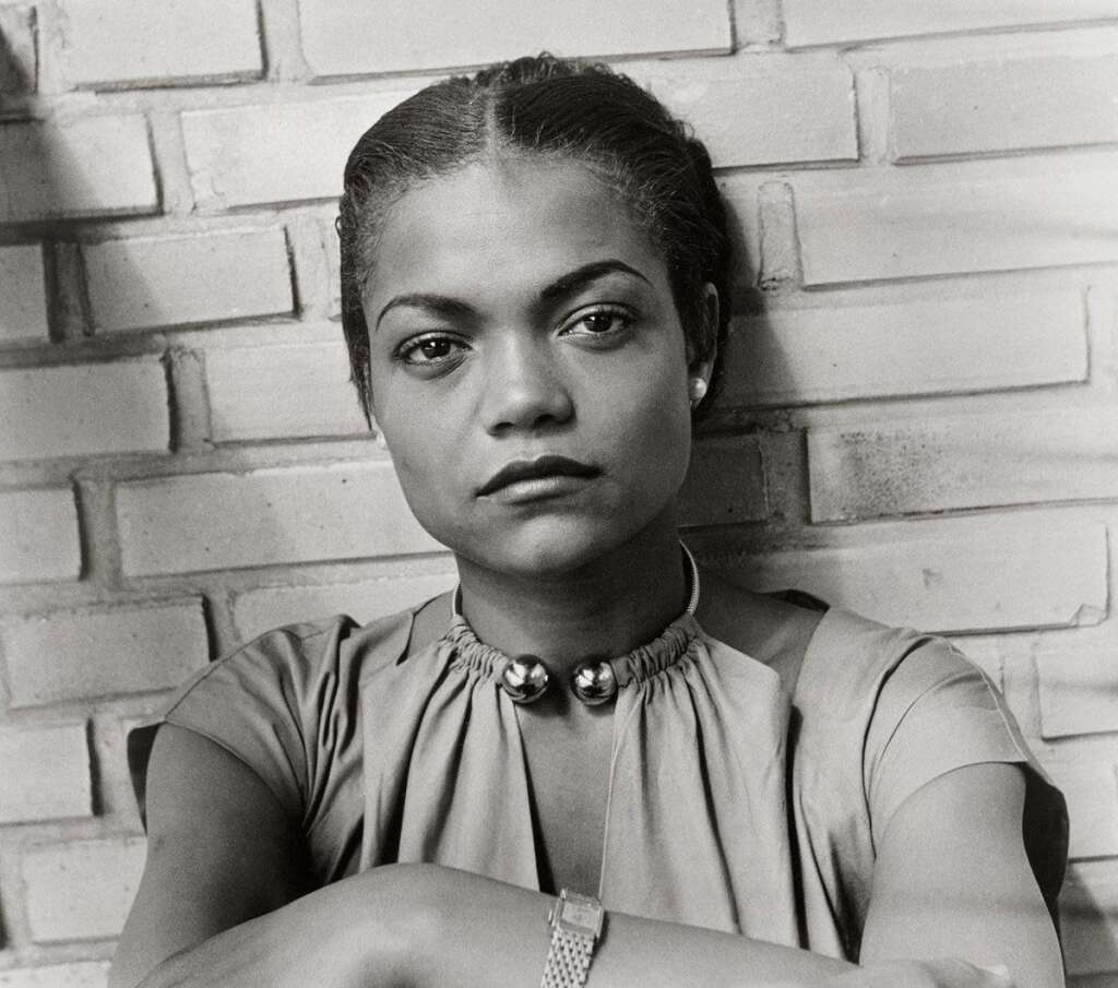 Black and white photograph of a woman with a serious expression. She has her hair pulled back and is leaning against a brick wall, wearing a light-colored dress with a high collar and two decorative buttons at the neck. She is looking directly at the camera.