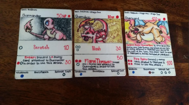 A trio of hand-drawn Pokémon cards featuring Charmander, Charmeleon, and Charizard. Each card displays the creature's unique abilities, hit points, and evolution stages, with illustrations and red text detailing their moves and attributes. They are placed on a wooden surface.