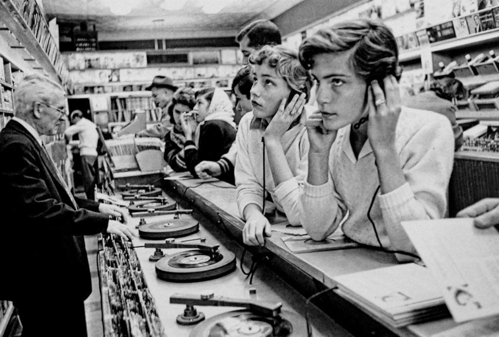 Black and white photo of people in a record store, listening to vinyl records on turntables. Two young women in the foreground are holding headphones to their ears. Shelves with album covers are visible in the background. An older man assists customers.