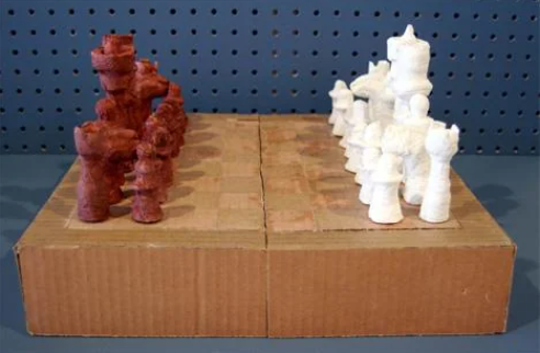 A chessboard made from a cardboard box with pieces crafted from clay or similar material. The pieces are roughly modeled, with one side colored brown and the other white, placed on a board marked with a grid of squares.