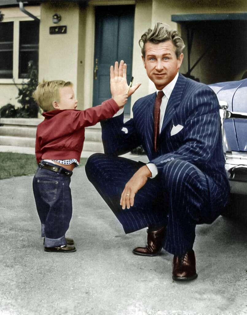 A young child gives a high-five to a man in a pinstripe suit, who is kneeling down. The man appears to be smiling, and they are standing outside in front of a house and a vintage car.