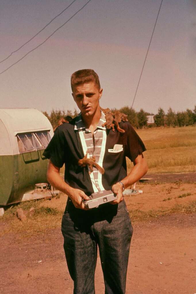 A young man with a serious expression stands outdoors, holding a small rectangular object. He wears a black shirt with light blue accents and has squirrels climbing on his shoulders and arms. Behind him, there is a green and white camper and a grassy field.