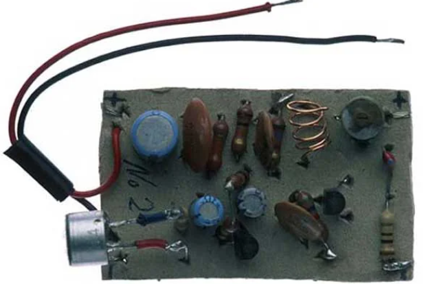 A small circuit board featuring various electronic components such as resistors, capacitors, diodes, transistors, and coils. Two wires, one red and one black, are connected to the board. The components are arranged in a compact, organized layout.