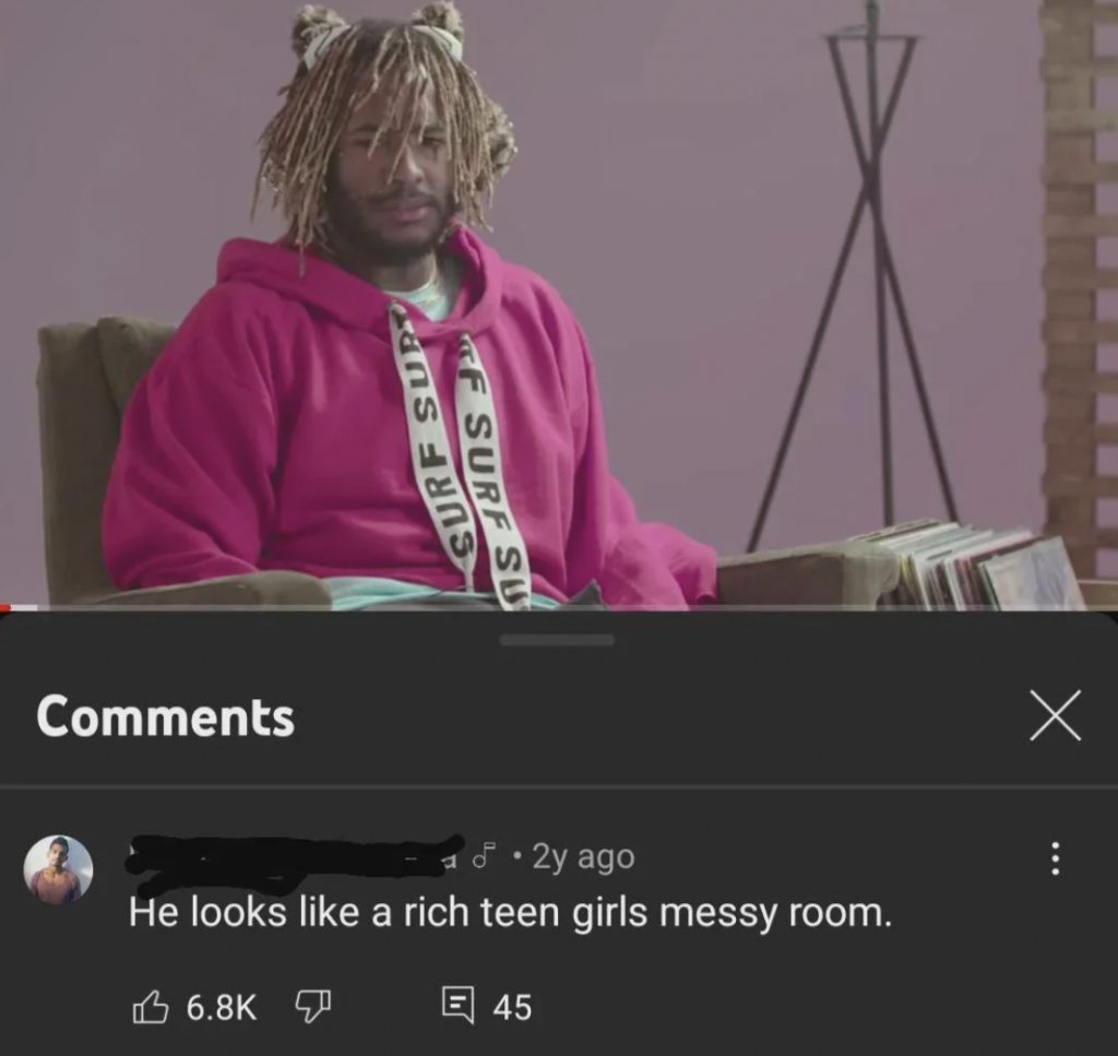 A person with light brown dreadlocks and wearing a pink hoodie with a white strap sits in a chair in a stylish, modern setting. Below the image, a YouTube comment reads, "He looks like a rich teen girl's messy room," with 6.8K likes and 45 replies.