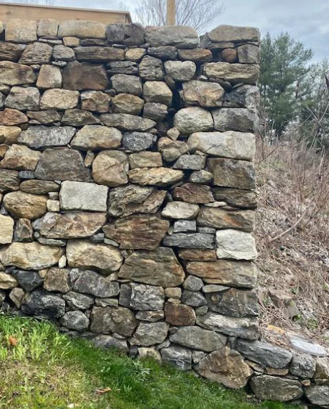 A weathered stone wall constructed from variously sized and shaped rocks sits on a patch of green grass. Behind the wall, there is sparse vegetation and a few trees under a cloudy sky.