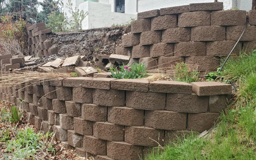 A retaining wall constructed from brown rectangular bricks with visible gaps and some bricks out of place. The wall supports a slope with exposed soil and a building in the background. Plants are growing in some of the spaces between the bricks.