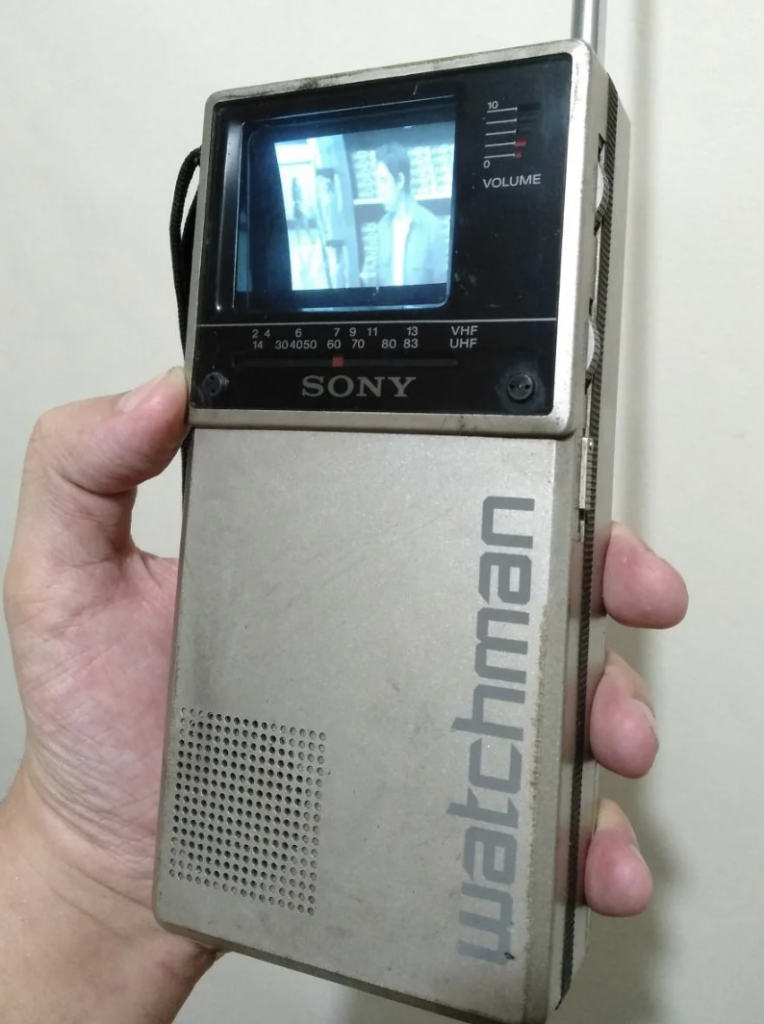 A person holds a vintage Sony Watchman portable TV. The device has a small screen showing a black and white image of people. The metallic body of the TV has a built-in speaker and volume controls on the side, with "watchman" written vertically.