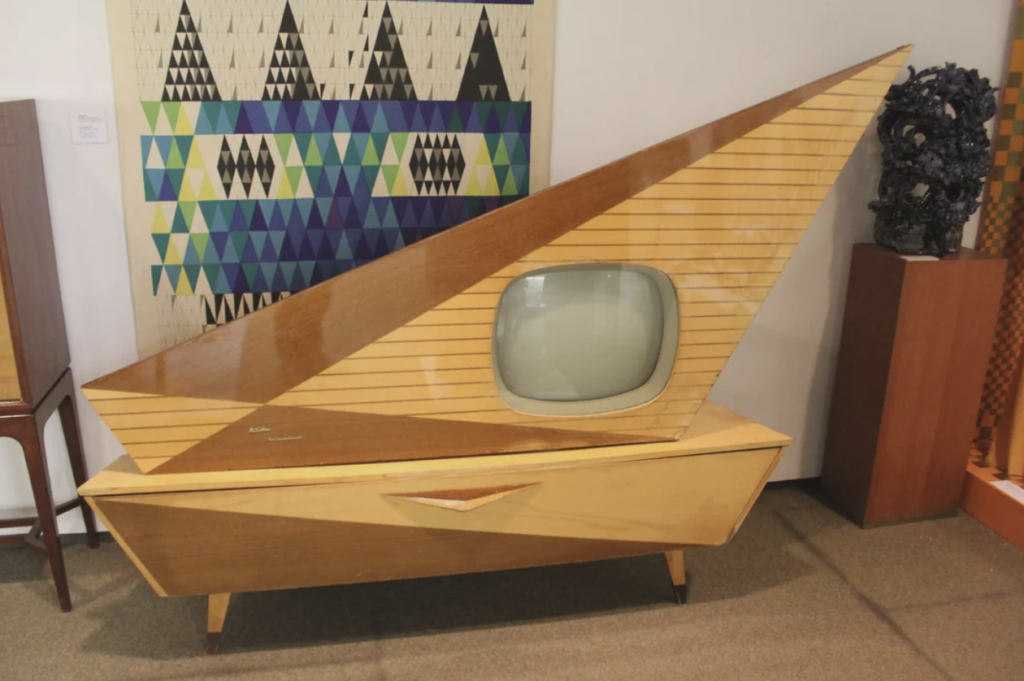 A vintage, mid-century modern television with a unique, triangular wooden frame and a rounded screen. It sits on an angular wooden cabinet, adorned with a striped pattern. A colorful geometric tapestry and various artworks are visible in the background.