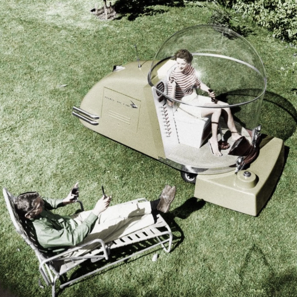 A woman with a futuristic, transparent dome on her head sits in a retro, pod-like vehicle, while a man lounges on a lawn chair nearby. They are in a grassy outdoor setting, both appearing relaxed and engaged in conversation.