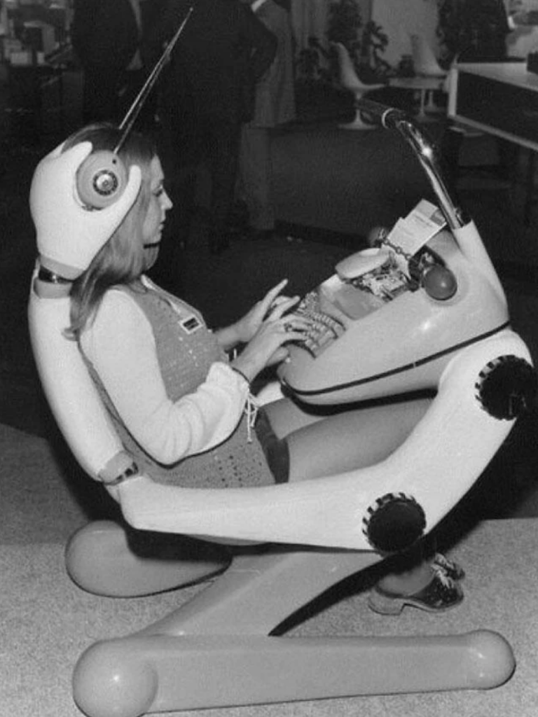 A woman sits in a futuristic, pod-like ergonomic chair that features a built-in typewriter and other controls. She is typing, and the chair is designed with large, articulated joints and a sleek, retro-futuristic aesthetic. The background is blurred and indistinct.