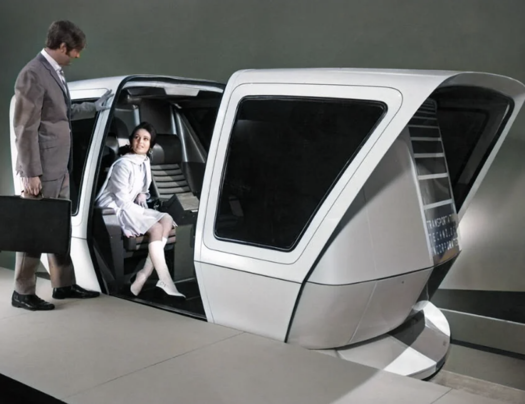 A man and a woman interact with a futuristic, pod-like vehicle. The woman, dressed in a light outfit, is seated inside the vehicle, while the man, holding a briefcase and wearing a suit, stands outside. The vehicle's sleek design features angular lines and large windows.