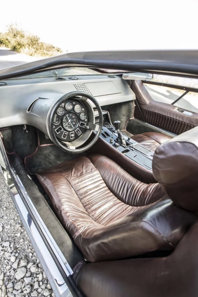 Interior view of a vintage car, showcasing a unique steering wheel with numerous gauges integrated into it, and a dashboard with various controls and buttons. The car has dark brown, leather-trimmed seats and panels, contributing to a retro-futuristic design.