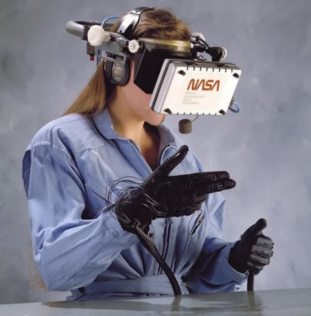 A person wearing a blue jumpsuit uses a vintage NASA virtual reality headset and wired gloves. The individual appears to be interacting with a virtual environment, against a neutral gray background.