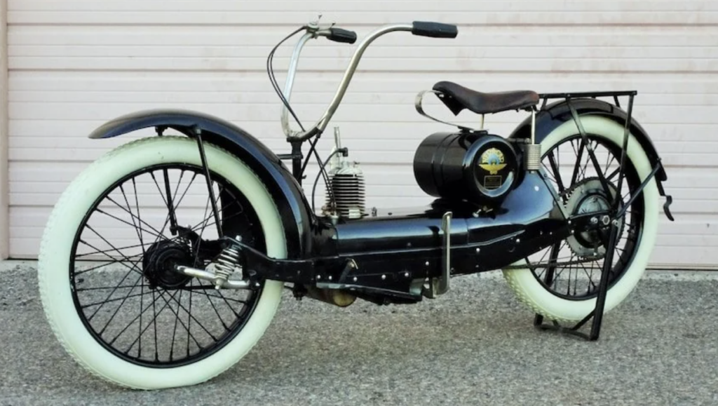 A vintage black motorcycle with large white tires is parked in front of a light pink garage door. The motorcycle features a prominent cylinder engine, exposed mechanical parts, and a brown leather saddle seat. The bike has a unique, minimalist frame design.