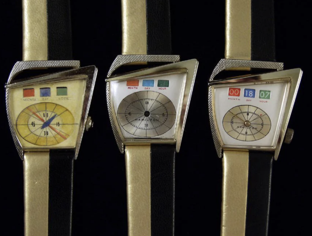 Three vintage triangular wristwatches with unique dials are displayed on black and beige straps. The left watch has a colorful dial with numbers and a slanted stripe. The center and right watches feature dater functions with numbers in color-tinted windows.