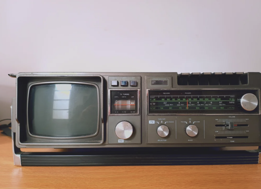 Vintage CRT television-radio combo on a wooden surface. The device features a small screen on the left, various dials, knobs, and switches in the center, and an AM/FM radio tuner with frequency markings on the right. The unit is set against a plain wall.