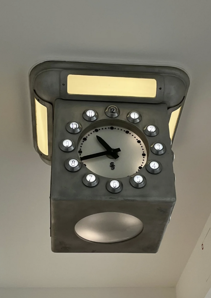 A clock designed to resemble a vintage rotary telephone dial. The clock face is situated in the center, with numbers arranged in a circular pattern around it, resembling a rotary dial. The design has a metallic finish and is mounted on the ceiling.