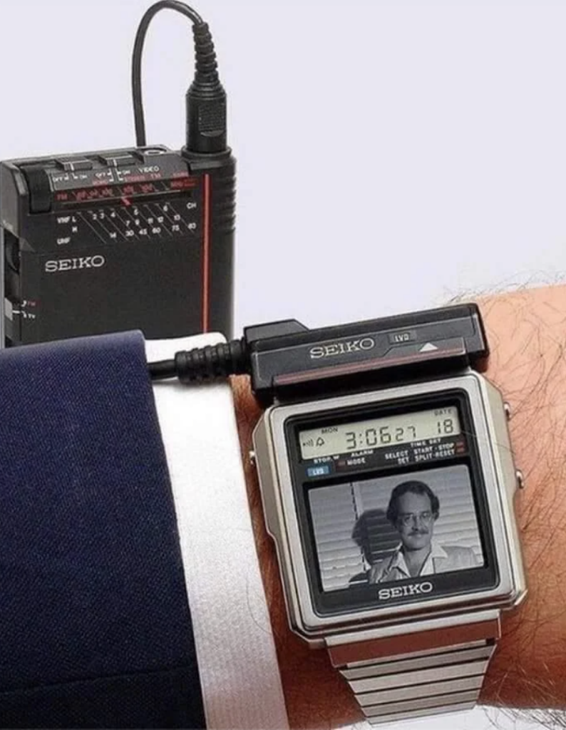 A person's wrist is shown wearing a retro Seiko TV watch, with a small screen displaying a black and white image. An attached external device, likely the television tuner, is also visible, with a cable connecting it to the watch. The person is wearing a suit.