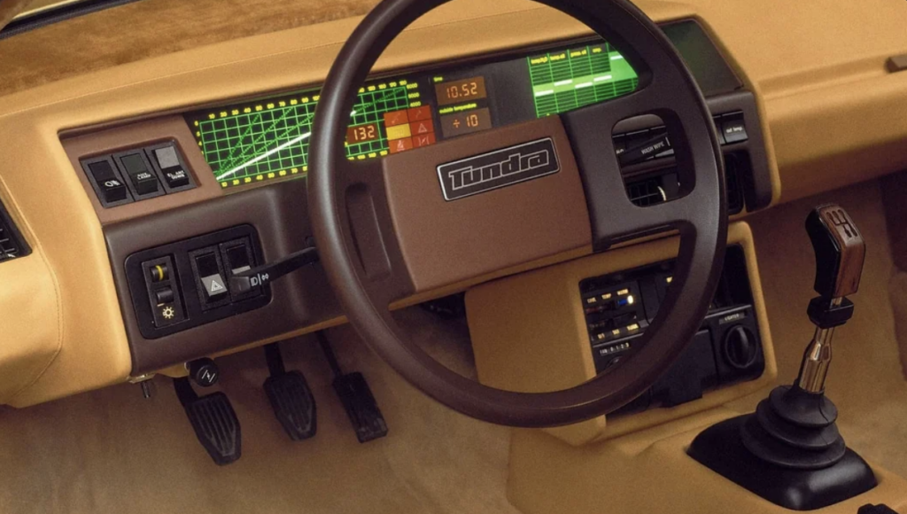 Vintage car dashboard featuring a digital display with graphs and speedometer, a steering wheel labeled "Tundra," manual gear shift lever, control buttons, foot pedals, and a beige interior. The design reflects a retro-futuristic style.