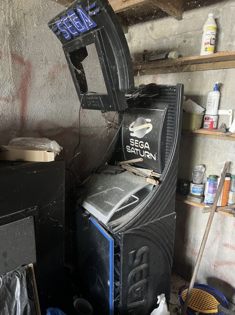 A neglected Sega Saturn arcade machine stands in a cluttered room with dusty shelves and cobwebs. The machine has a broken screen and a damaged marquee with the Sega logo. Various household items and tools are scattered around, underlining the machine's disuse.