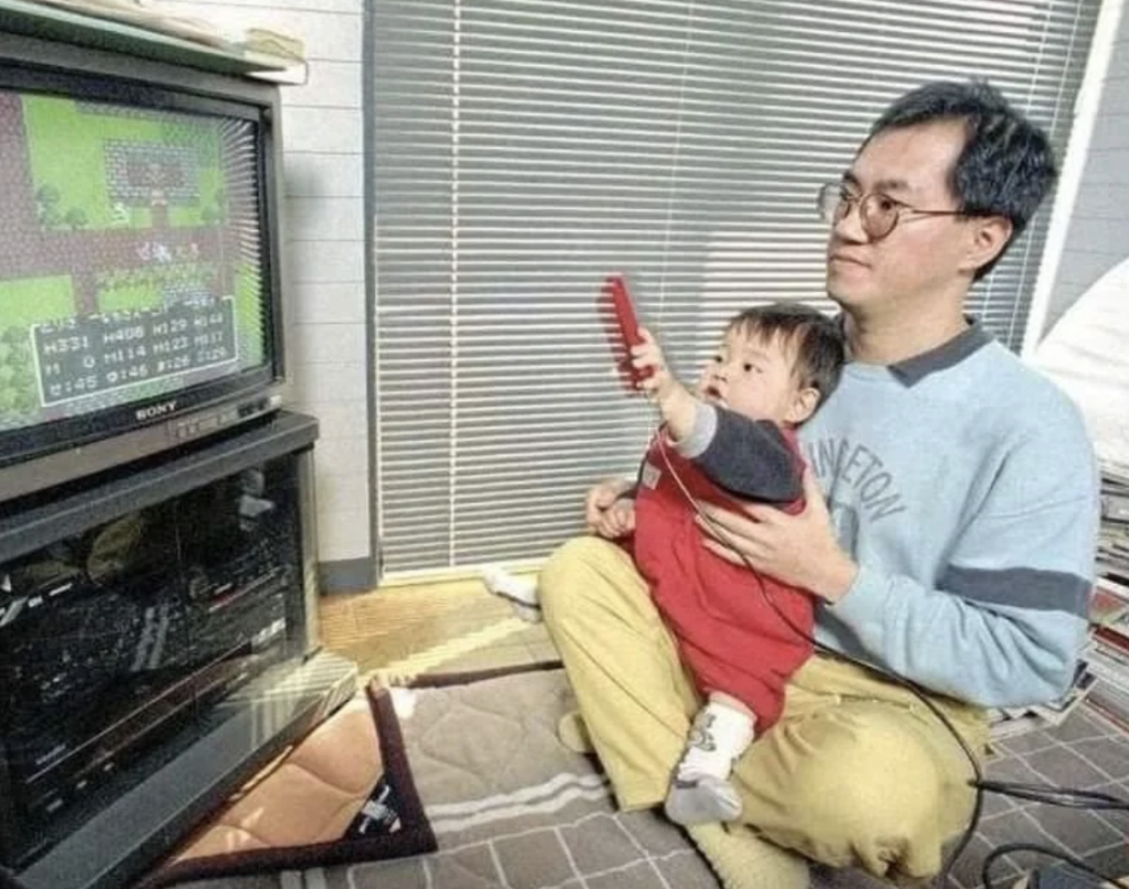 A man sits on the floor holding a young child who is reaching towards a television screen. The TV displays a vintage video game. The man is wearing glasses, a grey shirt, and yellow pants, while the child is dressed in a red and blue outfit.