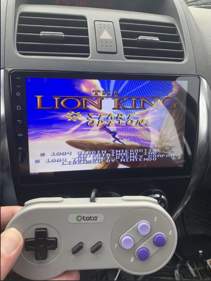 A person holds a retro gaming controller in front of a car's infotainment screen displaying the title screen of "The Lion King" video game. The controller has a white body with gray and purple buttons, and the infotainment screen is integrated into the car dashboard.