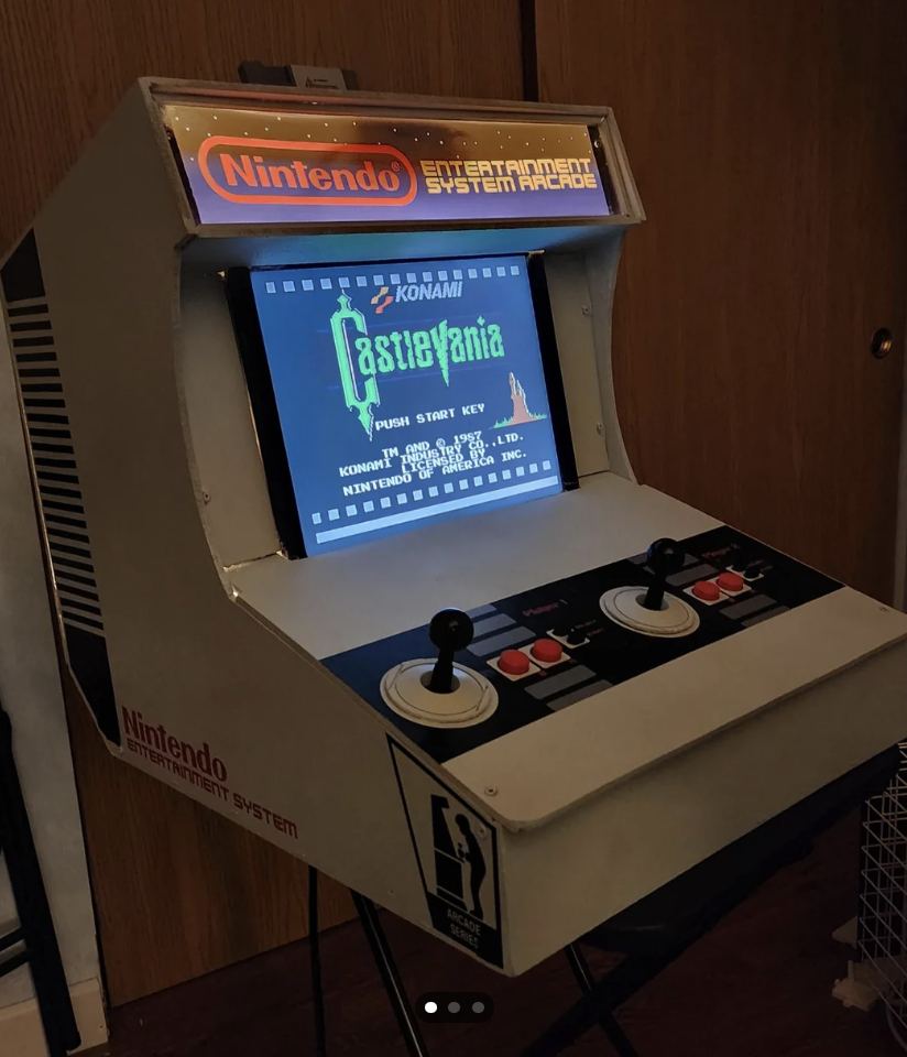 A retro-style Nintendo Entertainment System arcade machine displays the game screen for "Castlevania." The machine features joystick and button controls, and "Nintendo" is prominently labeled on its front and top panels. It sits in a room with wooden doors in the background.