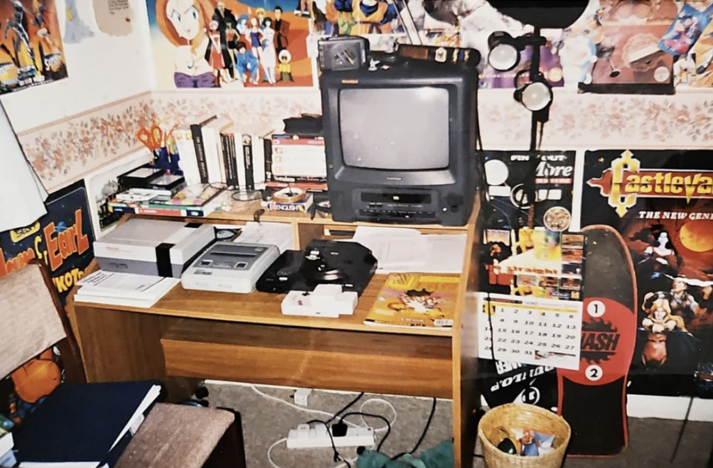 A cluttered desk in a room filled with various gaming and anime posters. The desk holds a CRT TV, video game consoles, books, and scattered gaming cartridges. Nearby objects include a calendar, a skateboard, office supplies, and a trash bin on the floor.