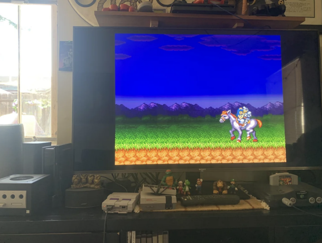 A living room with an old CRT television showing an animated character riding a horse in a pixelated landscape. Below the TV are various gaming consoles, including a Nintendo GameCube and a Super Nintendo Entertainment System (SNES).