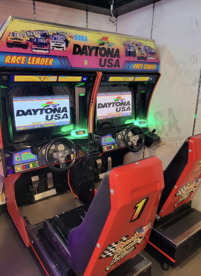 Two Sega Daytona USA arcade racing game machines side by side. Each machine has a steering wheel, seat, and pedals, with vibrant screens displaying "Daytona USA." The seats are red with checkered flag designs and a number "1" decal is visible on one seat.