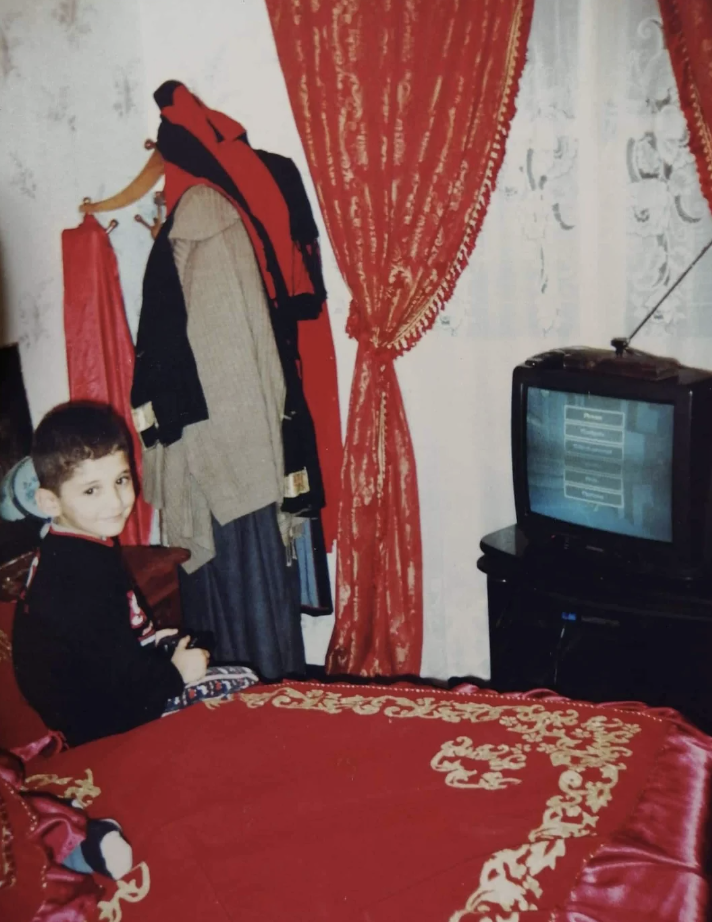 A young child sits on a bed covered with a red and gold bedspread, looking at the camera. Behind the child, elaborate red curtains frame a window, and a vintage television sits on a black stand showing a game. A coat rack holds hanging clothes next to the window.