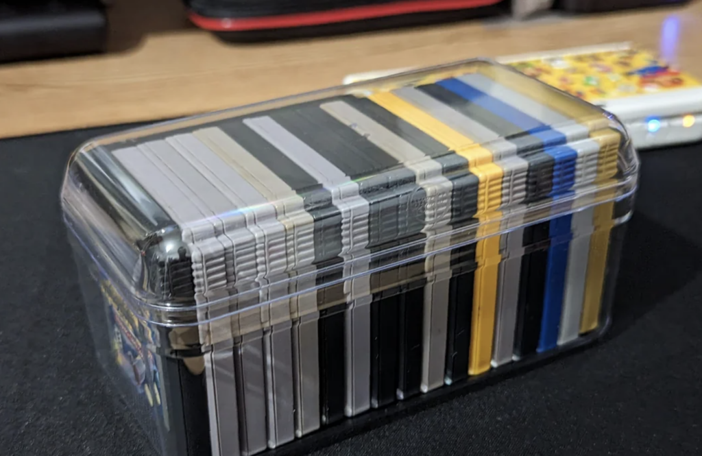 A plastic container holds a collection of card game decks, organized neatly within. The container is clear, showing various colored card sleeves inside, including black, yellow, and gray. The box is situated on a dark surface with a colorful item in the background.