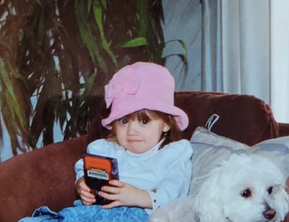 A young child wearing a pink hat and white shirt sits on a brown couch, holding a Game Boy. Beside the child, a small white dog rests on the couch. In the background, there are tall green houseplants.