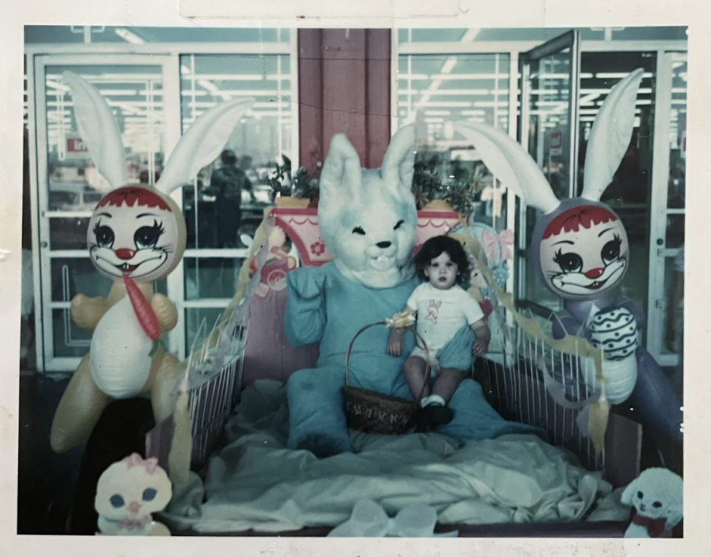 A child sits on the lap of a person in a large bunny costume, surrounded by two large, colorful bunny decorations with exaggerated facial expressions. They are in an indoor setting filled with bright, fluorescent lights. The scene has a festive, whimsical atmosphere.