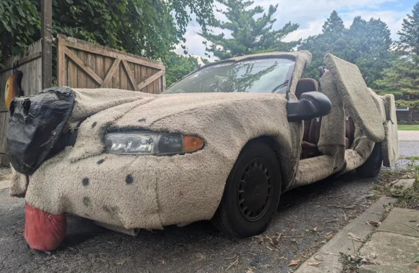 A car parked on a street that is entirely covered with shaggy, beige carpeting. The headlights and wheels are visible, and the car door is open. Trees and a wooden fence are in the background. The car has an unusual, furry appearance due to the carpeting.