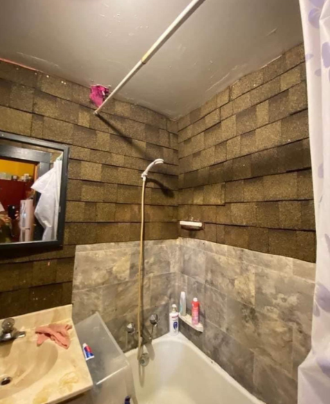 A small bathroom with a shower-tub combination. The shower walls are covered in brown shingles above a tiled lower section. Various toiletries are on the edge of the tub and sink. A shower curtain with a light pattern hangs partially open.