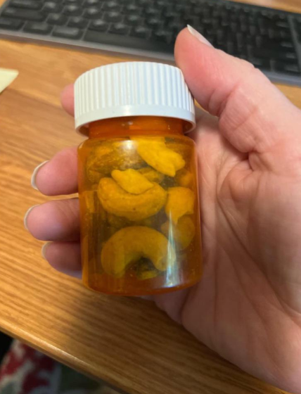 A hand holding a small, amber-colored prescription bottle filled with cashew nuts instead of medication. A keyboard and part of a desk are visible in the background.