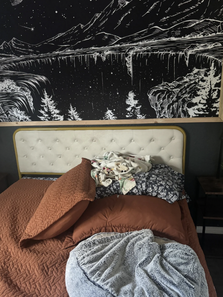 A bedroom with a beige tufted headboard, adorned with multiple pillows and blankets in various colors and patterns. Above the bed is a large, monochromatic wall mural depicting a nighttime forest scene with trees, cliffs, and a starry sky.