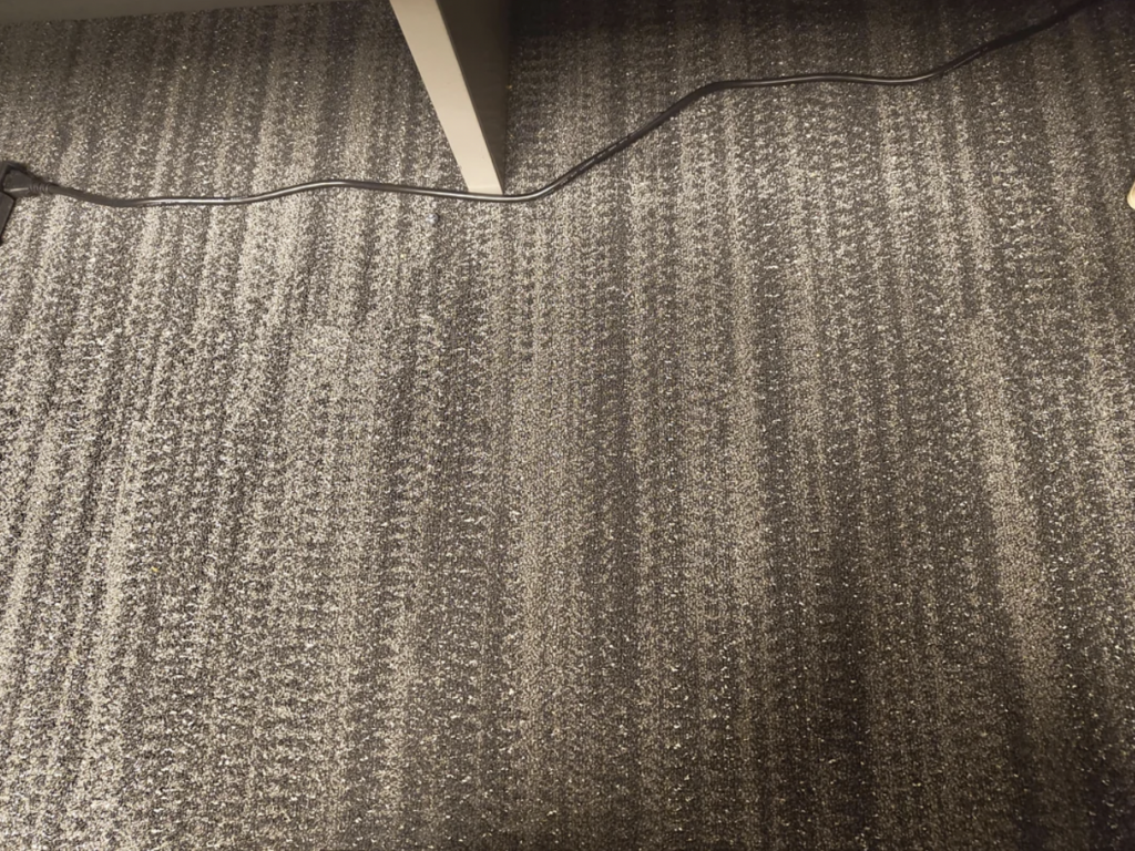 A patterned carpet with horizontal and vertical lines in shades of gray. An electrical cord runs across the middle of the floor beneath what appears to be the leg of a table or desk on the left side.