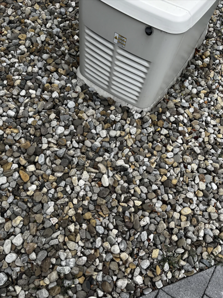 A gray utility box with a white top is situated outdoors, surrounded by small, multicolored rocks. The area appears to be part of a landscaped section with more rocks extending outward from the box, adjacent to a paved surface on the right.