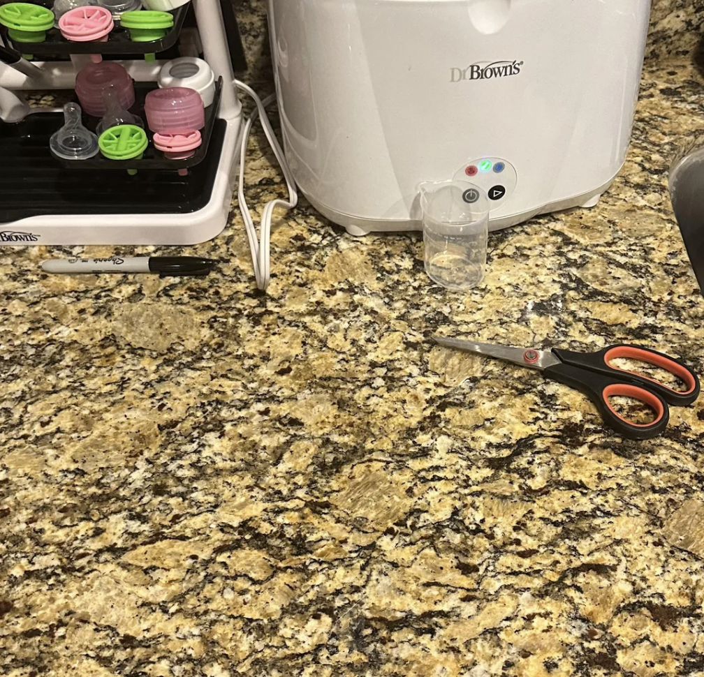 A kitchen counter with a baby bottle drying rack holding bottles and accessories, a Dr. Brown's bottle sterilizer, a black marker, a clear measuring cup, and a pair of scissors with red handles. The countertop is granite with a mix of brown, tan, and black colors.